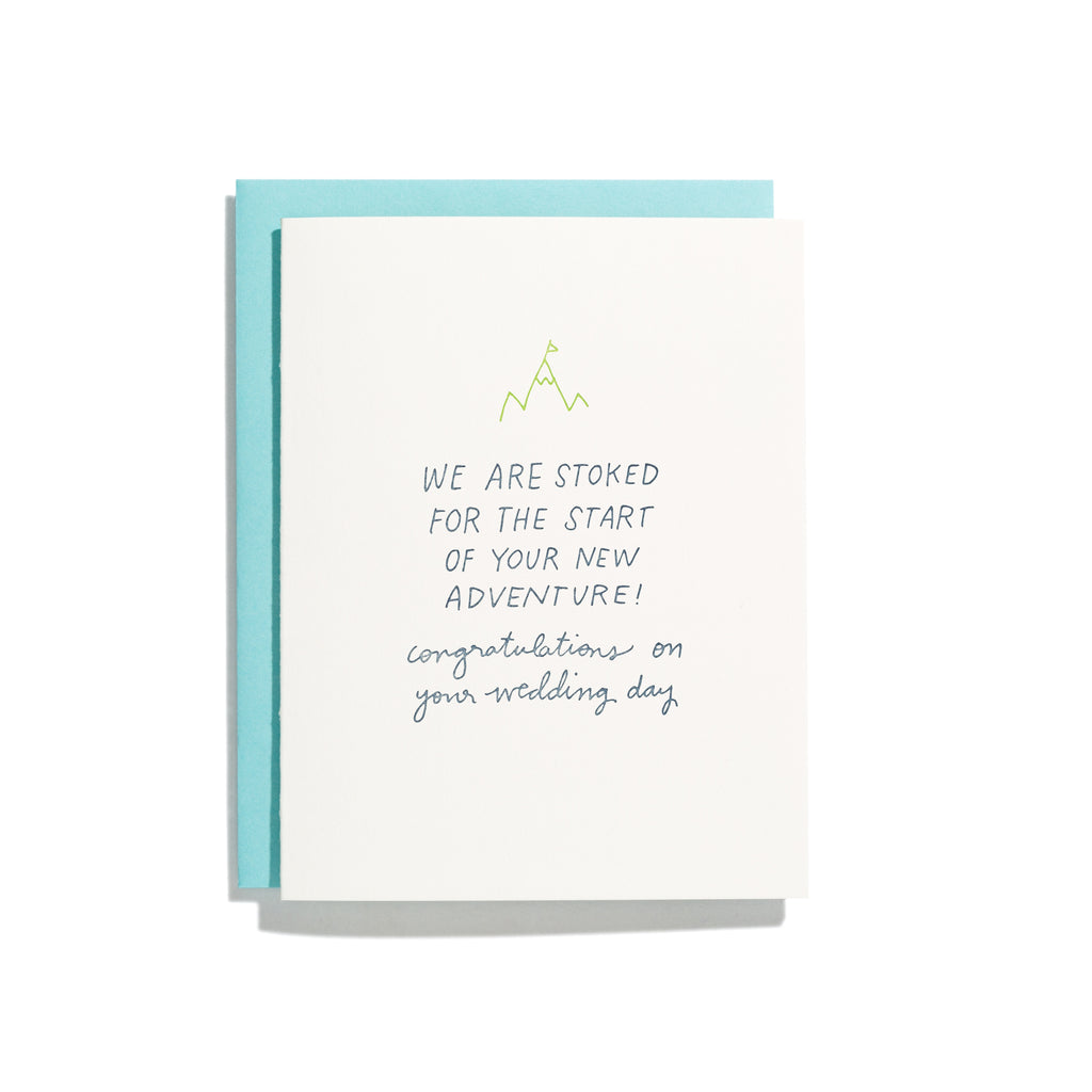 White card with blue text saying, “We are stoked for the start of your new adventure! Congratulations on your wedding day”. Image of a green mountaintop above text. A blue envelope is included.