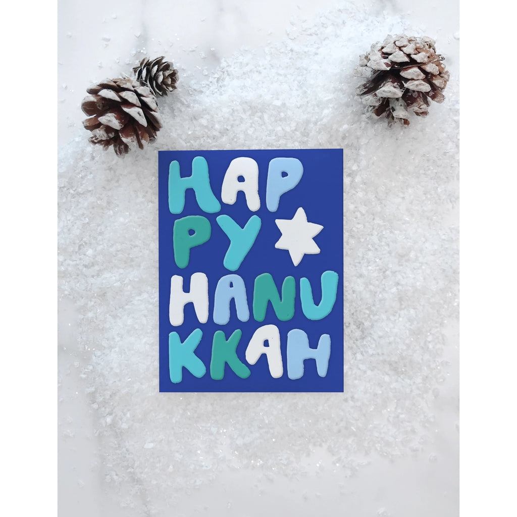 Dark blue card with light blue and white text saying, “Happy Hanukkah”. Image of a white star. An envelope is included.