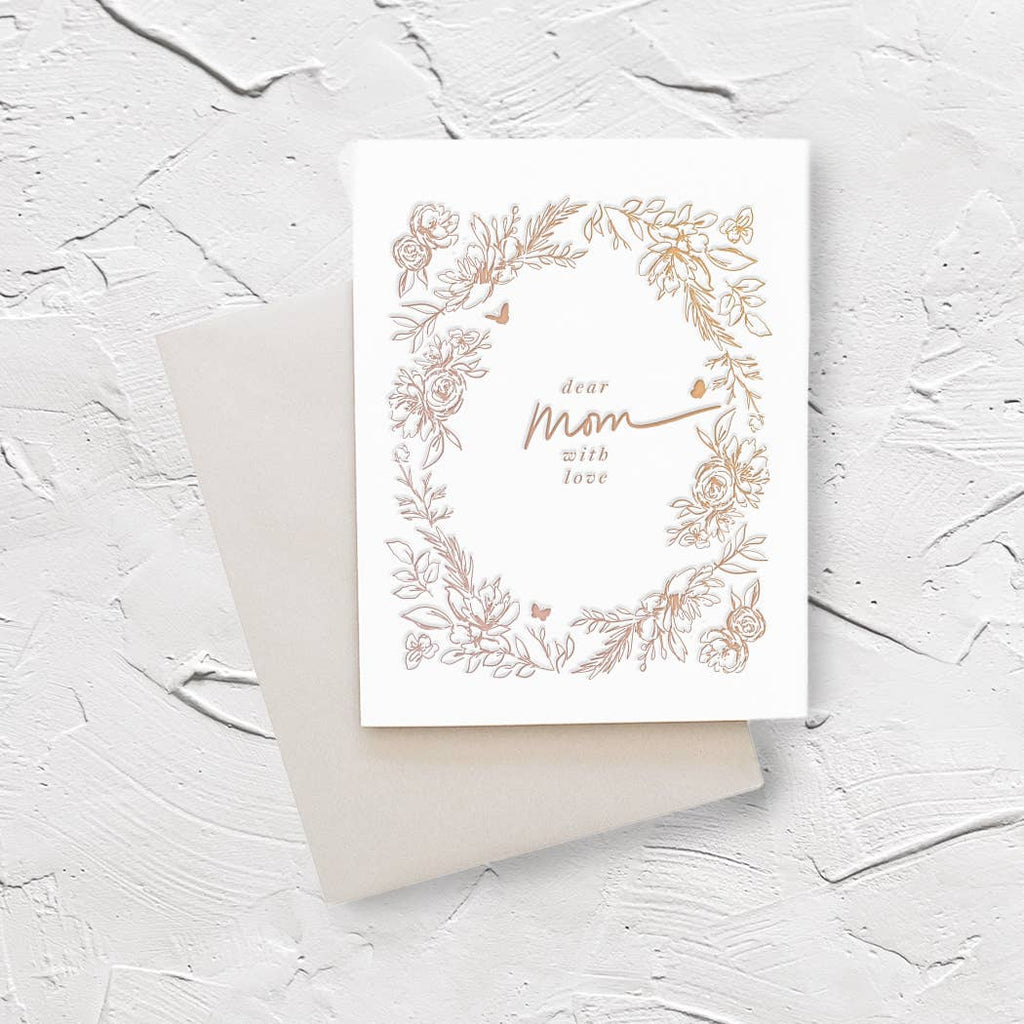 Ivory background with tan images of flowers, leaves and butterflies. Text in middle says in tan says, “Dear Mom with love”. A silver envelope is included.   