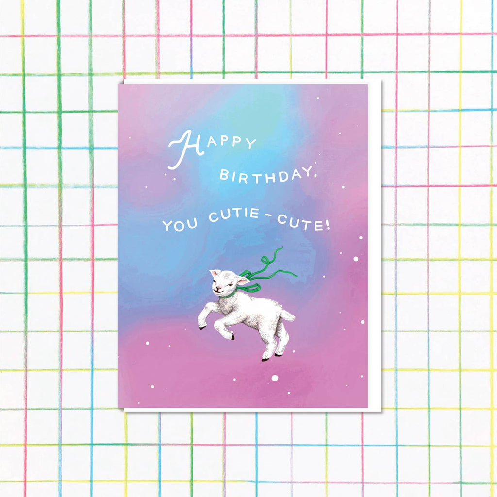 Purple and blue background with random white dots and image of a small white lamb wearing a green ribbon around its neck. White text says, “Happy Birthday, You Cutie-cute!”. A white envelope is included. 