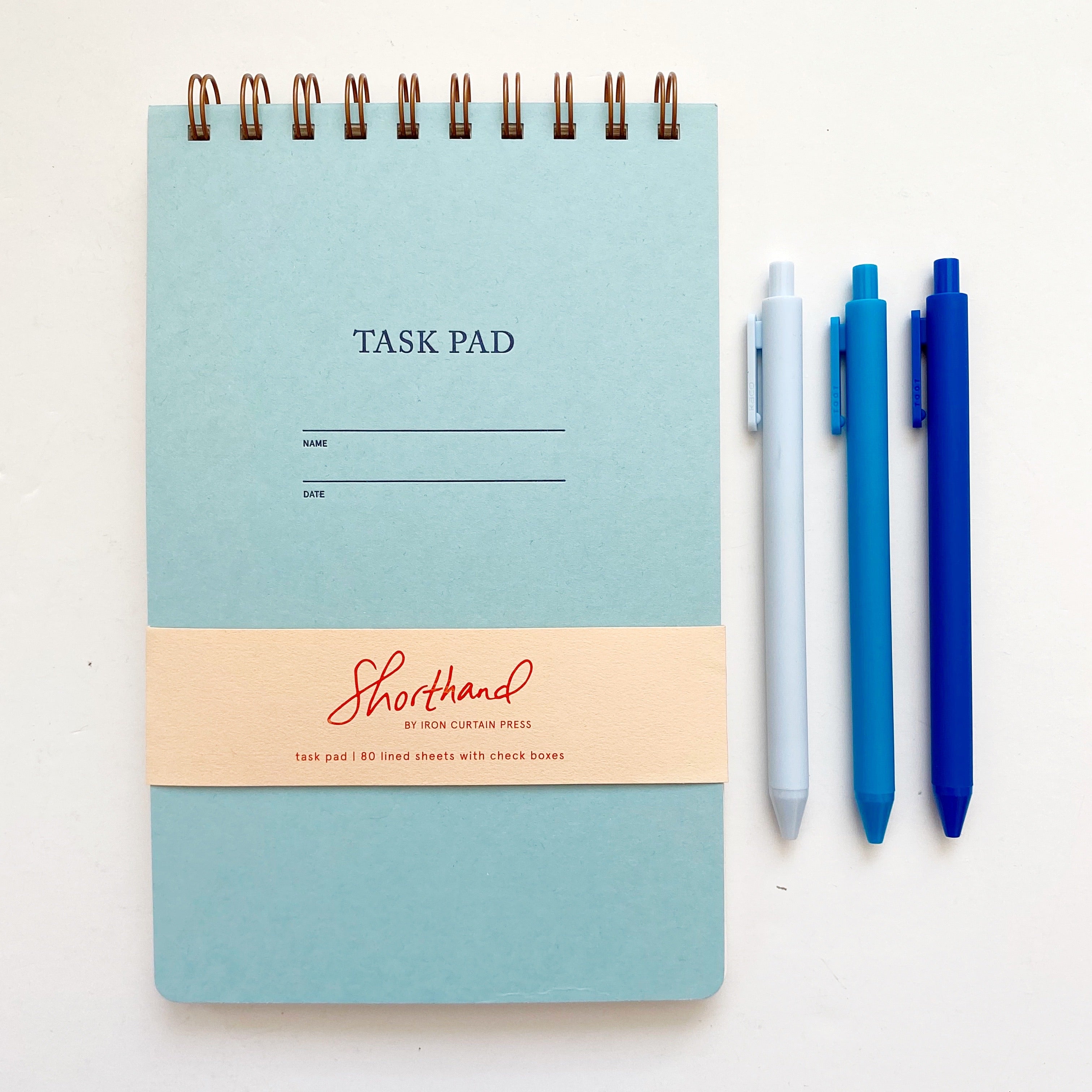 Image of pool blue with letter pressed text says, “Task pad”. “Name” and “Date” with lines for writing. Coiled binding on top with three blue pens.