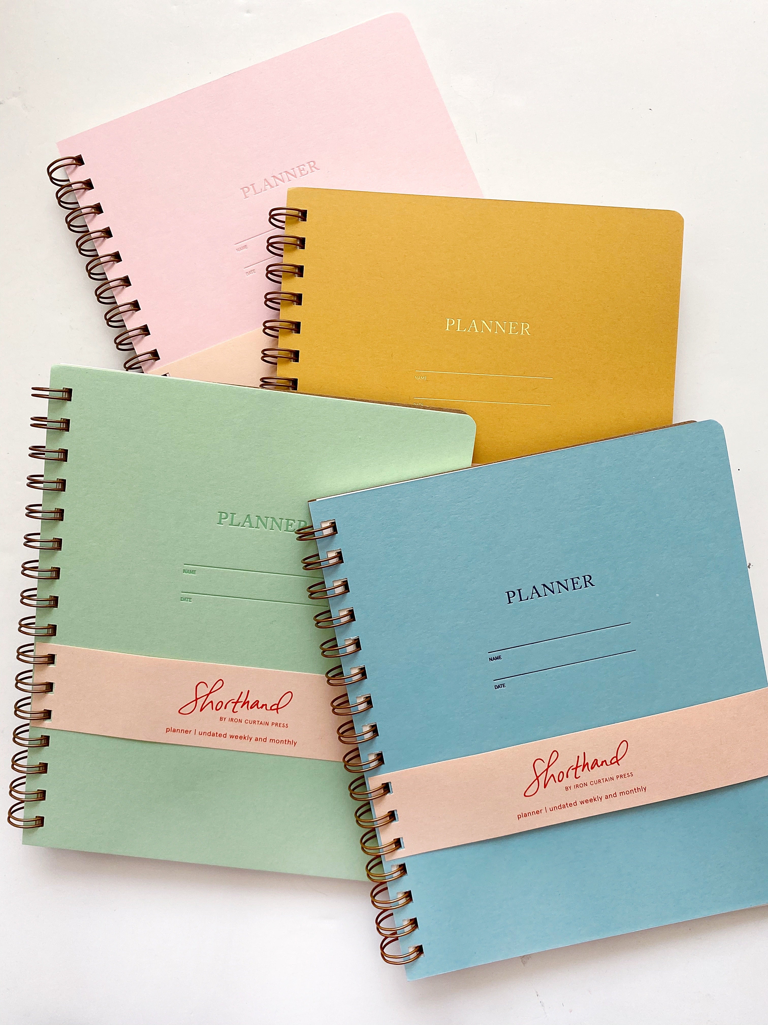 Image of planner covers in light pink, mustard, pool, and mint with side coiled binding. Letter pressed text says, “Planner”, “name” and “date” with lines for writing,